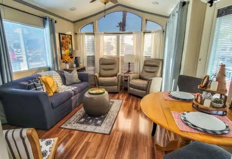 A cozy living room with large windows in a 1998 Chariot Eagle features a sofa, two armchairs, and a round coffee table. Adjacent is a dining area with a round table set for two, perfect for intimate meals. Wood flooring adds warmth throughout the space.