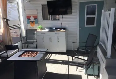 Covered patio of this 1998 Chariot Eagle mobile home features a wall-mounted TV, white sideboard, square fire pit table, black chairs, teal door, and decorative wall art. Light streaming through curtains creates captivating shadows on the floor.