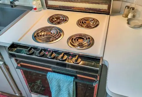 A vintage 1998 Chariot Eagle stove with four coil burners graces the white surface. A blue towel hangs on the oven door handle, evoking a sense of nostalgia. The kitchen countertop and sink are partially visible in this charming RV setting.
