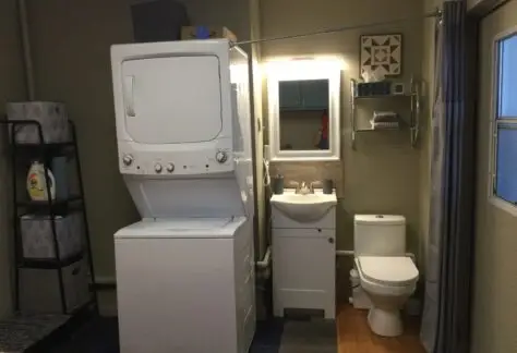 The compact bathroom in the 1998 Chariot Eagle model features a stacked washer and dryer, small sink, toilet, and efficient shelving. A mirror with a light hangs above the sink, while a curtain partially obscures a window to the right.