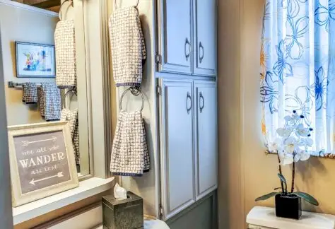 A bathroom in a Chariot Eagle mobile home features a sink, mirror, and wall cabinet. A framed quote adorns the wall, while a floral curtain graces the window. An elegant potted orchid sits next to the toilet, adding a touch of nature to this charming 1998 model space.
