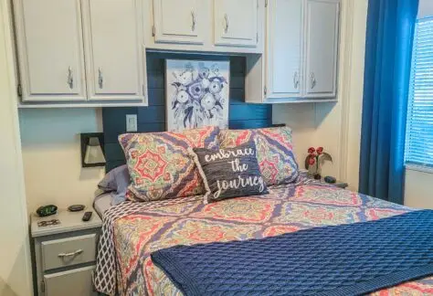 A cozy bedroom in the 1998 Chariot Eagle mobile home boasts a bed with colorful patterned bedding and a decorative pillow, flanked by cabinets and a small nightstand. A window with dark curtains completes the inviting space.