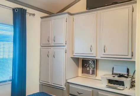 The cozy bedroom features gray cabinets, a wall-mounted TV, and a window with elegant curtains. A bed with a patterned cover adds comfort, while below the TV, a printer rests on the shelf—a perfect modern touch reminiscent of classic 1998 Chariot Eagle design.