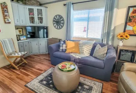Cozy living room with a 1998 vibe, featuring a blue sofa, patterned cushions, and a round coffee table with snacks. A striped chair complements the Chariot Eagle decor, while sunflowers in a vase add charm near the window with curtains. A wall-mounted clock and TV complete the scene.