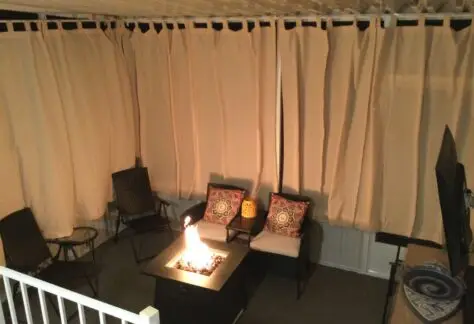 The outdoor patio, reminiscent of a 1998 Chariot Eagle's charm, features wicker chairs with colorful cushions and a fire pit elegantly surrounded by beige curtains.