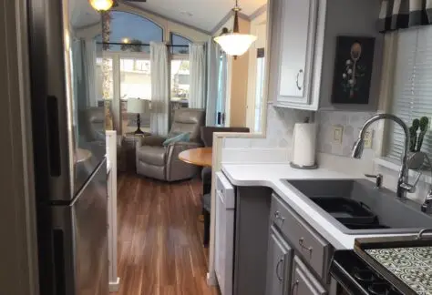 In this cozy Chariot Eagle kitchen, sleek gray countertops and stainless steel appliances harmonize with white cabinets and a decorative backsplash. Through the doorway, a living area with plush recliners echoes the charm of 1998 design.