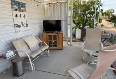 Patio on Lot 1 features a small TV on a wooden stand, two cushioned chairs, a small round table, and string lights overhead. A Cavco touch is evident with desert plants in the background and an artistic wall hanging adding charm to the space.