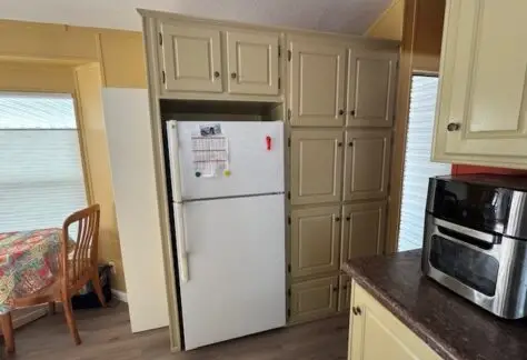 A kitchen with a white refrigerator surrounded by beige cabinets, reminiscent of a classic 1998 design. A small oven sits on the counter to the right. On the left, a table with a colorful tablecloth adds charm to this Cavco-inspired space.