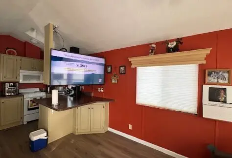 The living room, painted in rich red, features sleek cabinets and a mounted TV displaying text about Lot 1. A nostalgic 1998 calendar hangs nearby, while a window with a shade and various treasures from Cavco adorn the shelves, adding unique charm to the space.