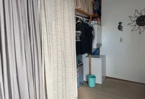 A cozy nook in the 1998 Cavco, Lot 1 features a curtain revealing a closet area where clothes hang neatly. In the corner, a washing machine and dryer are stacked. Above a turquoise bin, a decorative sun adds charm to the space.