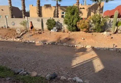 Dry landscaped backyard with rock and gravel ground, sparse plants, and desert-themed decorations. A plaster wall offers a hint of serenity with trees in the background casting intricate shadows. This scene perfectly complements the timeless appeal of a 1998 Cavco design on Lot 1.