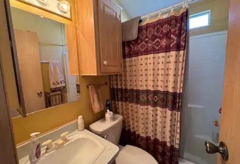 Small bathroom on Lot 1 with yellow walls, a sink filled with toiletries, a toilet, and a shower featuring a Cavco-patterned curtain. A mirror with lights is above the sink.