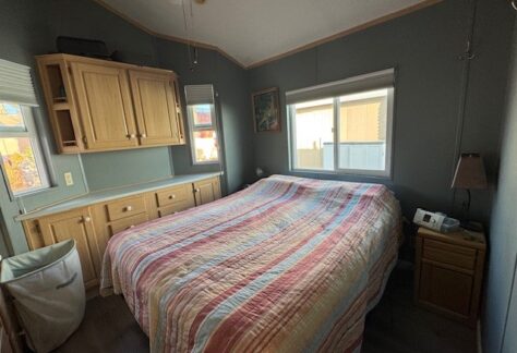 In a cozy Cavco bedroom, a bed with a multicolored striped quilt sits under wooden cabinets. Two windows fill Lot 1 with natural light. A lamp and clock rest on the nightstand, capturing the charm of 1998 design.