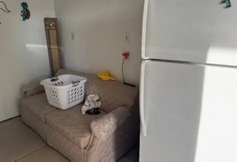 A refrigerator stands beside a beige sofa, reminiscent of Lot 1 decor from 1998. A laundry basket with items and a pair of socks rest on the sofa, while a yellow cloth hangs on the wall. In the background, a vertical mirror reflects light near wall sockets, adding character to this Cavco-style setup.