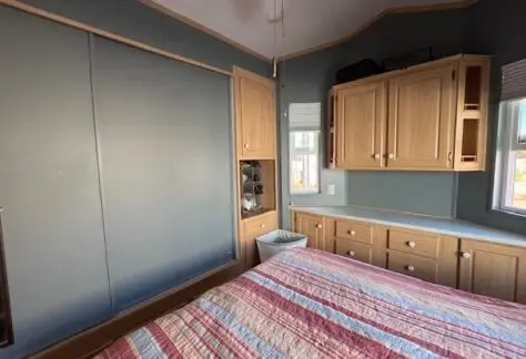 This 1998 Cavco bedroom features wood-tone cabinetry, a striped bedspread, and minimal decor. With blue walls, closed curtains, and natural light peeking through the windows of Lot 1, it offers a cozy retreat.