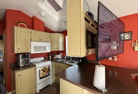 The 1998 Cavco kitchen features red walls and beige cabinets, complemented by a gas stove, microwave, coffee maker, and mounted TV. Skylight above and paper towel holder on the counter add to the charm of Lot 1's cozy culinary space.