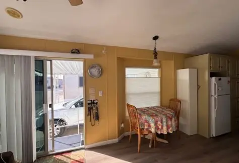 A cozy, small dining area featuring a round table draped in a colorful cloth and two wooden chairs. The room, reminiscent of a classic 1998 Cavco design, includes a sliding glass door leading outside and showcases a wall clock alongside the refrigerator.