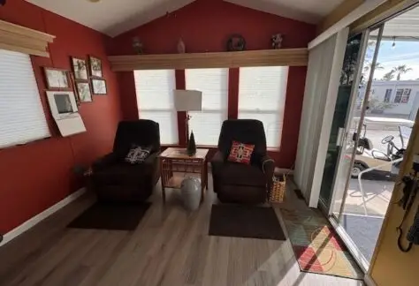 A cozy living room featuring two brown armchairs, a lamp, a side table, and a wicker basket adds charm to the 1998 Cavco design. Red walls adorned with framed pictures complement the space, while a sliding glass door to the right invites natural light into Lot 1.