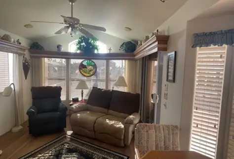 A cozy Cavco living room on Lot 123 features a reclining sofa, armchair, ceiling fan, and large windows with blinds. A stained glass decoration hangs in the window above a patterned rug on the floor.