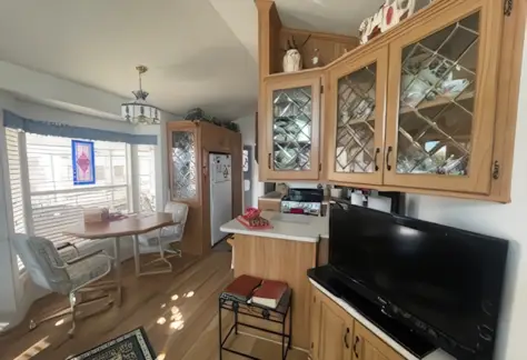 A cozy kitchen in Lot 123 features Cavco wooden cabinets and a small TV. A round table with chairs sits by a window adorned with blinds and stained glass, while a rug adds warmth to the light wood floor.