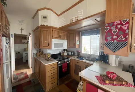 A cozy kitchen with wooden cabinets and white countertops, featuring a red theme. Decorative towels add charm, while appliances glisten. In the background of Lot 123, a glimpse of a hallway and household items hints at the home’s inviting layout.
