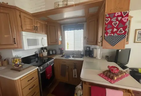 Located in Lot 123, this Cavco-designed compact kitchen features wooden cabinets, white countertops, an oven, microwave, and a range of other appliances. A window with blinds sits above the sink, complemented by a decorative towel hanging nearby.