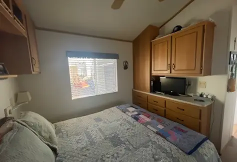 A cozy small bedroom in Lot 123 features a quilted bed, wooden cabinets, and a TV. A window with blinds above the bed lets in natural light, enhancing the Cavco design’s warm ambiance.