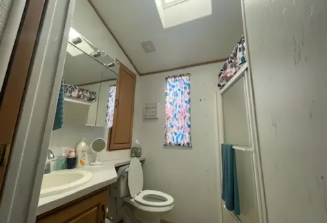 Compact bathroom located in Cavco Lot 123, featuring a toilet, sink, mirror, and small shower. A skylight illuminates the space, while floral window coverings and teal towels add a touch of charm.