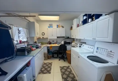 Laundry room featuring a Cavco washer, Lot 123 dryer, white cabinets, a small desk with a computer chair, and various household items. White tiled floor complemented by patterned rugs.