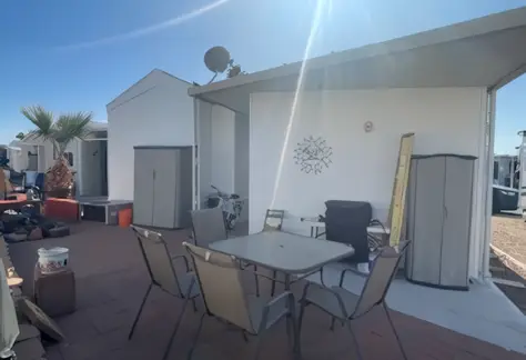 Outdoor patio with a glass table and several chairs, adjacent to a white Cavco building. A palm tree sways gently next to storage cabinets and various items scattered around Lot 123. Sunlight shines overhead, creating a warm, inviting atmosphere.