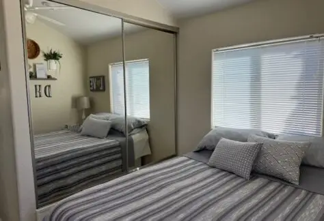 A cozy Chariot Eagle bedroom features a striped bedspread, decorative pillows, and mirrored closet doors reflecting a window with blinds. The walls display framed decor, complemented by a lamp on the bedside table.