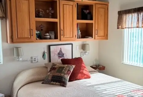 Bedroom with a neatly made bed, decorative pillows, and a patterned blanket. Wooden cabinets with plants and knick-knacks are above the bed. A painting is hung on the wall. Sunlight enters through a window.