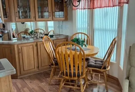 A cozy dining area with a round wooden table, four chairs, a cabinet with glass doors, and a chandelier. The room has wooden flooring and a large window with vertical blinds and red curtains.