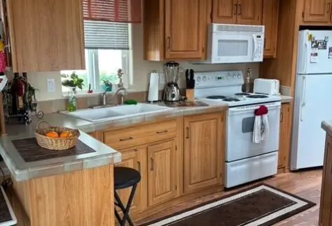 A wooden kitchen with a stove, microwave, and refrigerator. A fruit basket and blender sit on the counter. A brown rug runs along the floor.