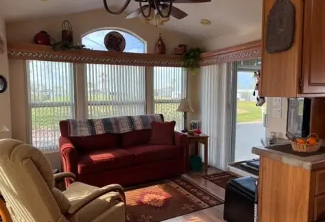 Cozy living room with a red sofa, beige armchair, and decorative items on shelves. Sunlight filters through large windows with vertical blinds, highlighting a patterned rug on the floor.