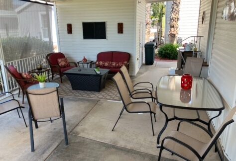 Outdoor patio with red cushioned seating, glass tables, chairs, and potted plants. Covered area with wall-mounted lights, a sink, and palm trees in the background.