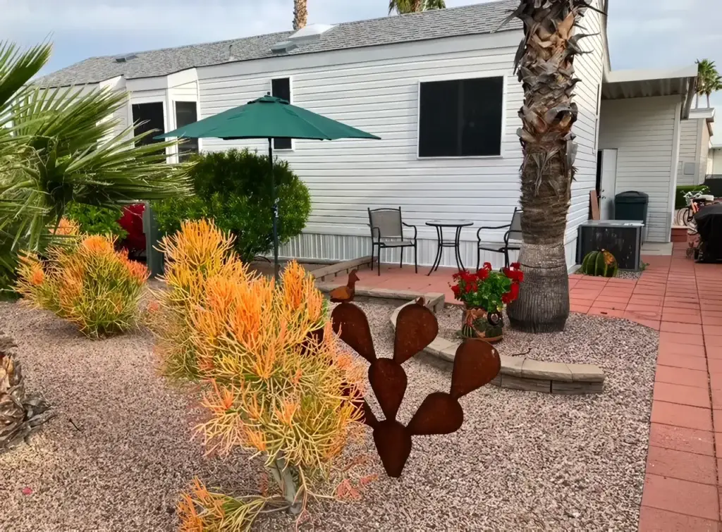 A mobile home with a small patio area. Patio features a table with two chairs under a green umbrella. There's desert landscaping with plants, a palm tree, and a metal cactus sculpture.