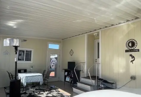 The indoor patio of Lot 430 features charming yellow walls and a dining table adorned with a floral tablecloth. A TV and decorative wall hangings complete the cozy ambiance, while a heater rests discreetly beneath the metal ceiling. Built by Cavco in 2013, this space blends style and comfort effortlessly.