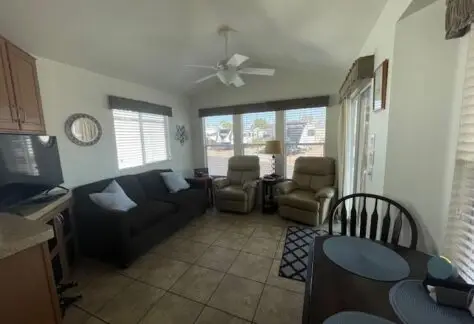 This cozy living room features a Cavco charm with a brown sofa, two beige armchairs, and a flat-screen TV. A small dining table sits nearby. Large windows with blinds let in natural light, highlighting the tiled flooring of Lot 430. A ceiling fan gracefully spins overhead.