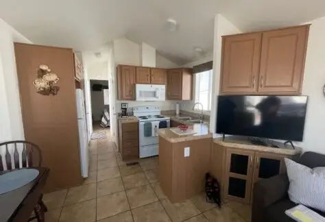 Compact kitchen and living area with wooden cabinets, white appliances, a countertop, and a wall-mounted TV. Tan floor tiles and a dining table are visible in this 2013 Cavco design at Lot 430.