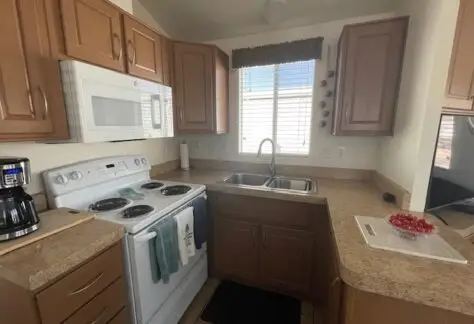 Compact kitchen in Lot 430 featuring a white stove, microwave, and coffee maker on the countertop. Brown cabinets, double sink, window with blinds, and a small bowl of cherry tomatoes complete this 2013 Cavco design.