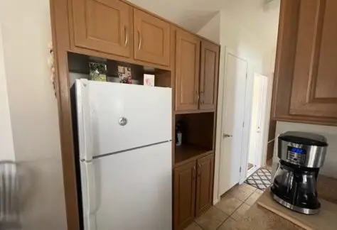 Wooden kitchen cabinets with a white refrigerator and a coffee maker on the counter create a cozy vibe in this Cavco home. A hallway elegantly leads to Lot 430, with its door offering a glimpse of inviting spaces beyond.