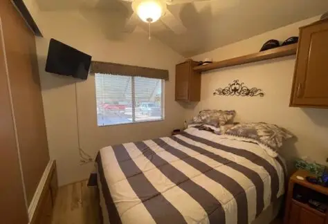 This cozy Cavco bedroom, nestled in Lot 430, features a double bed with striped bedding, a wall-mounted TV, and ceiling fan. A window with blinds brightens the space while a shelf holds hats and decorations.