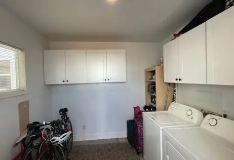 Located in Lot 430, this Cavco-designed laundry room boasts a 2013 look with its modern washing machine and dryer on the right. Bicycles rest on the left beneath bright white cabinets mounted on the wall, all illuminated by natural light streaming through a window.