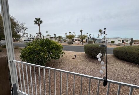From the porch of Lot 309, you can enjoy the view of a gravel yard framed by swaying palm trees with mobile homes lining the opposite street. A charming hanging decoration swings gently in the foreground on the right, adding a personal touch to this serene 2015 Cavco setting.