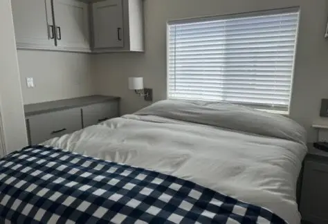 A bedroom with a neatly made bed featuring white linens and a blue checkered throw. The room has closed cabinets, a window with blinds, and a wall-mounted light on the left, perfectly styled to be the Champion 2023 of tranquil spaces.