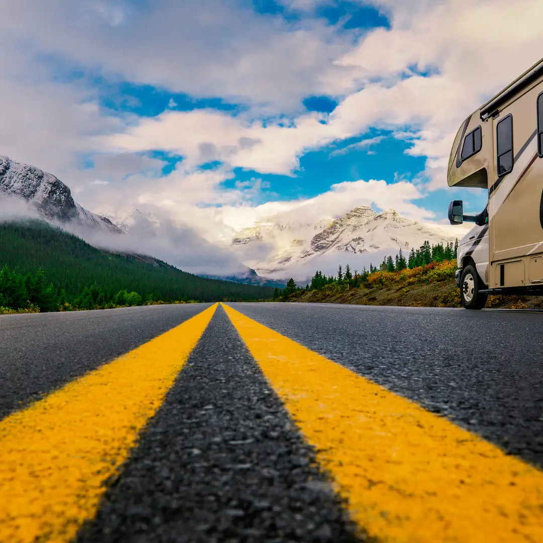 A camper van is parked by a road with a double yellow line, leading towards the snow-kissed mountains and cloud-draped peaks of Durango, all under a clear blue sky—a perfect start for spring adventures and outdoor fun.