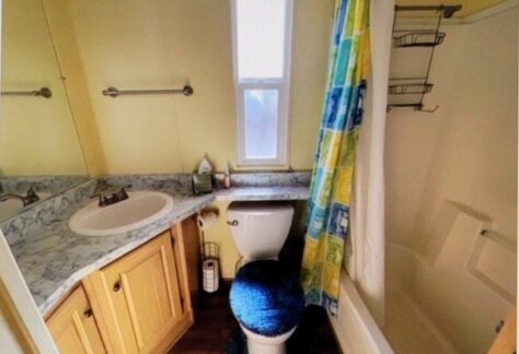 Charming small bathroom in Cavco's Lot 25, featuring a sink, toilet, and bathtub with wooden cabinets. A mirror enhances space alongside a colorful shower curtain, while shelves in the 2000-era design provide practical storage.