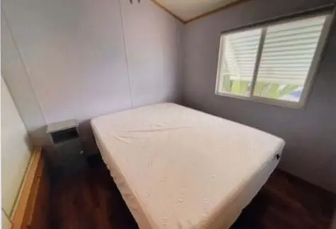 Small bedroom in Lot 25 features a white mattress on a bed frame, accompanied by a single nightstand. A window with closed blinds is positioned on the right wall, adding to the cozy ambiance of this 2000 Cavco model.
