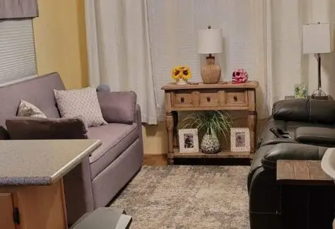 The living room on Lot 25 features a gray Cavco couch, armchair, curtains, and a ceiling fan. Decorative flowers adorn a wooden table over a plush rug on the hardwood floor.